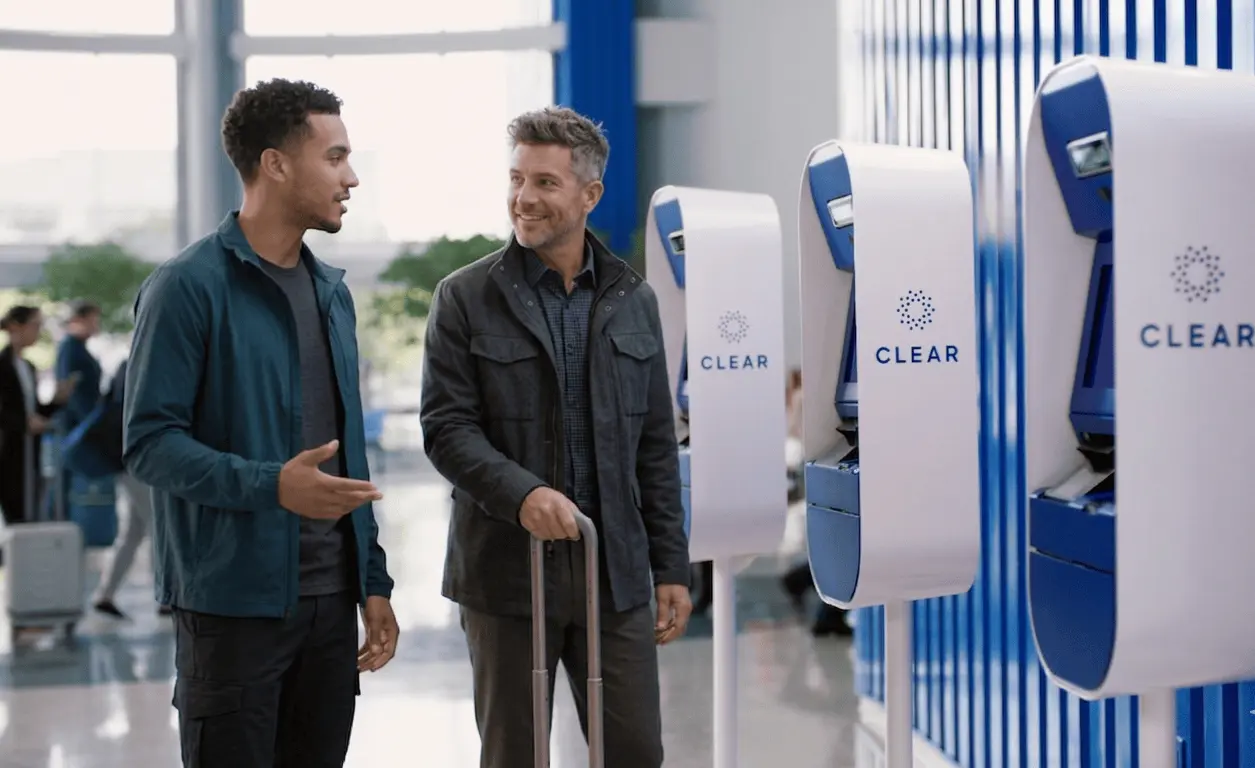 Two men standing at CLEAR kiosk at the airport smiling since they got CLEAR for free.